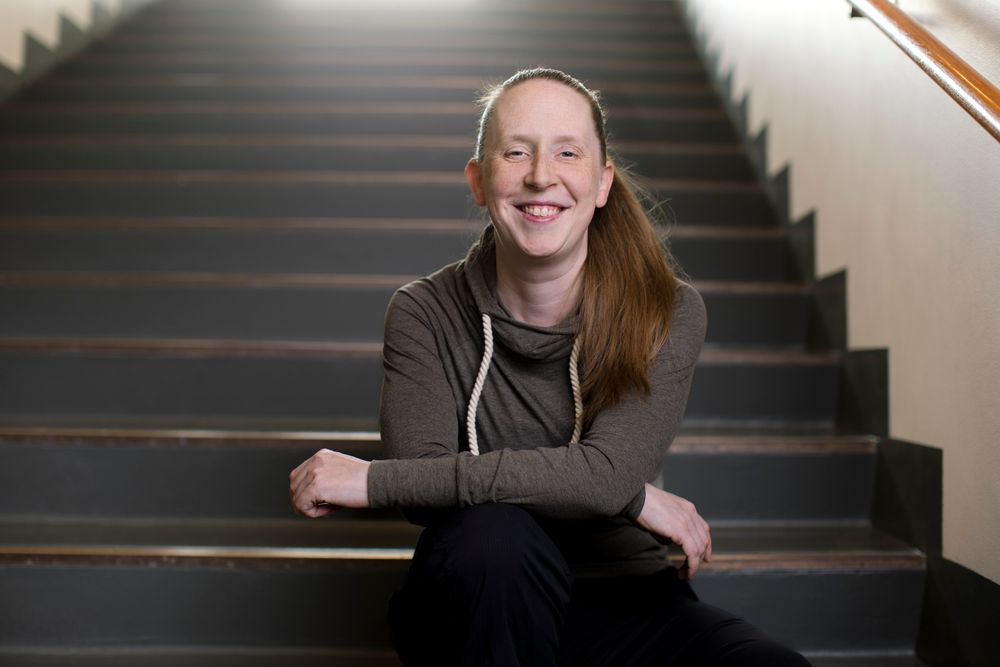 Een persoon zit op een brede trap in een binnenruimte met zachte verlichting. De persoon draagt een donkere, lange‑mouwtop met een capuchon en heeft een lange staart haar over één schouder. De armen zijn ontspannen gekruist over de knieën, en de trap loopt omhoog tot buiten beeld.