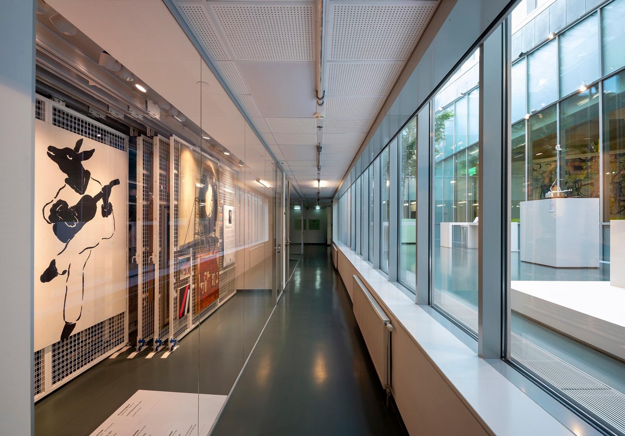De gang van het kunstdepot, met links achter glas de rekken waar schilderijen hangen en rechts het atrium met zicht op presentatiemeubels. 