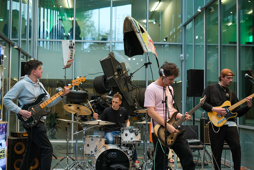 Bandje met vier jonge mannen treden op in het Atrium. Drie jongens spelen gitaar en een drumt. Op de achtergrond is een installatie van Jean Tinguely te zien.