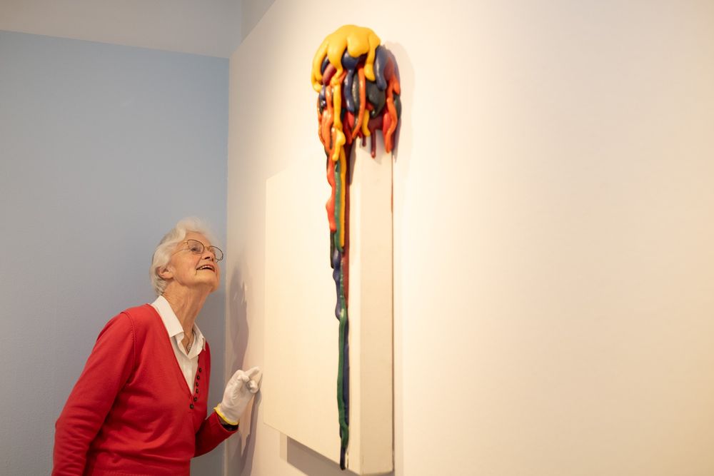 Een oudere vrouw in een rode trui kijkt naar een kunstwerk, voornamelijk wit, met aan de bovenkant een stapel kleuren die naar beneden druipen.