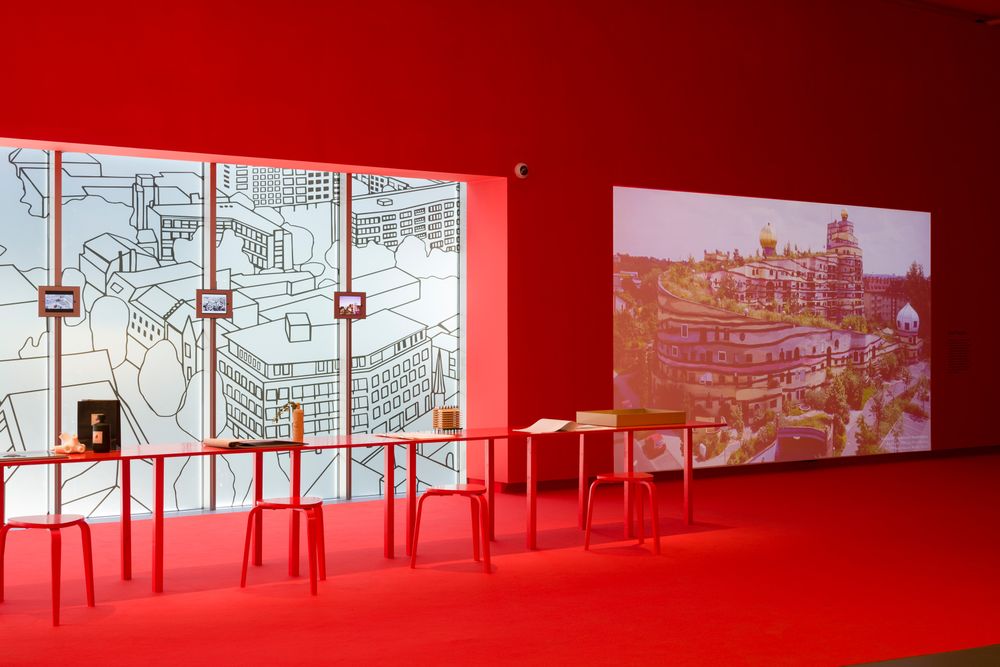Tentoonstellingsruimte in rood verlichte zaal met tafels, krukken en een grote projectie op de wand; op de achtergrond een raam met lijntekening van een stad.