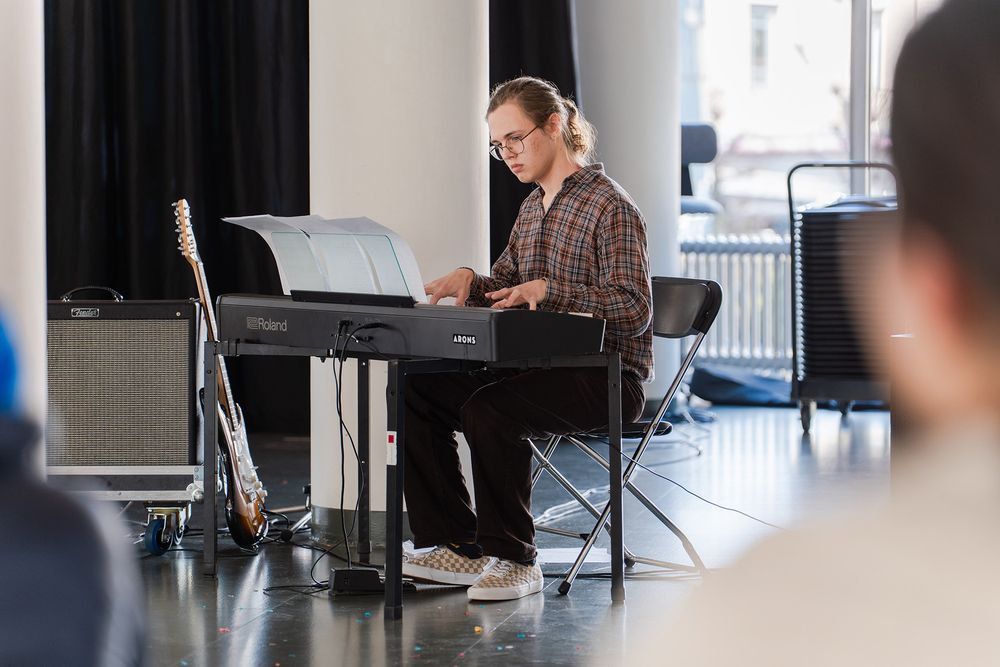 Persoon speelt op een elektrisch keyboard in een lichte ruimte, met bladmuziek voor zich. Links naast het keyboard staat een gitaar en versterker.