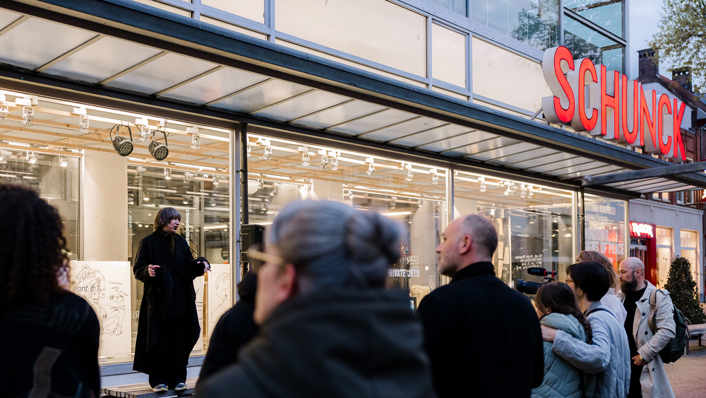 Bezoekers kijken naar Alevtina Kakhidze die buiten op een bankje voor de Vitrine van SCHUNCK de bezoekers toespreekt.