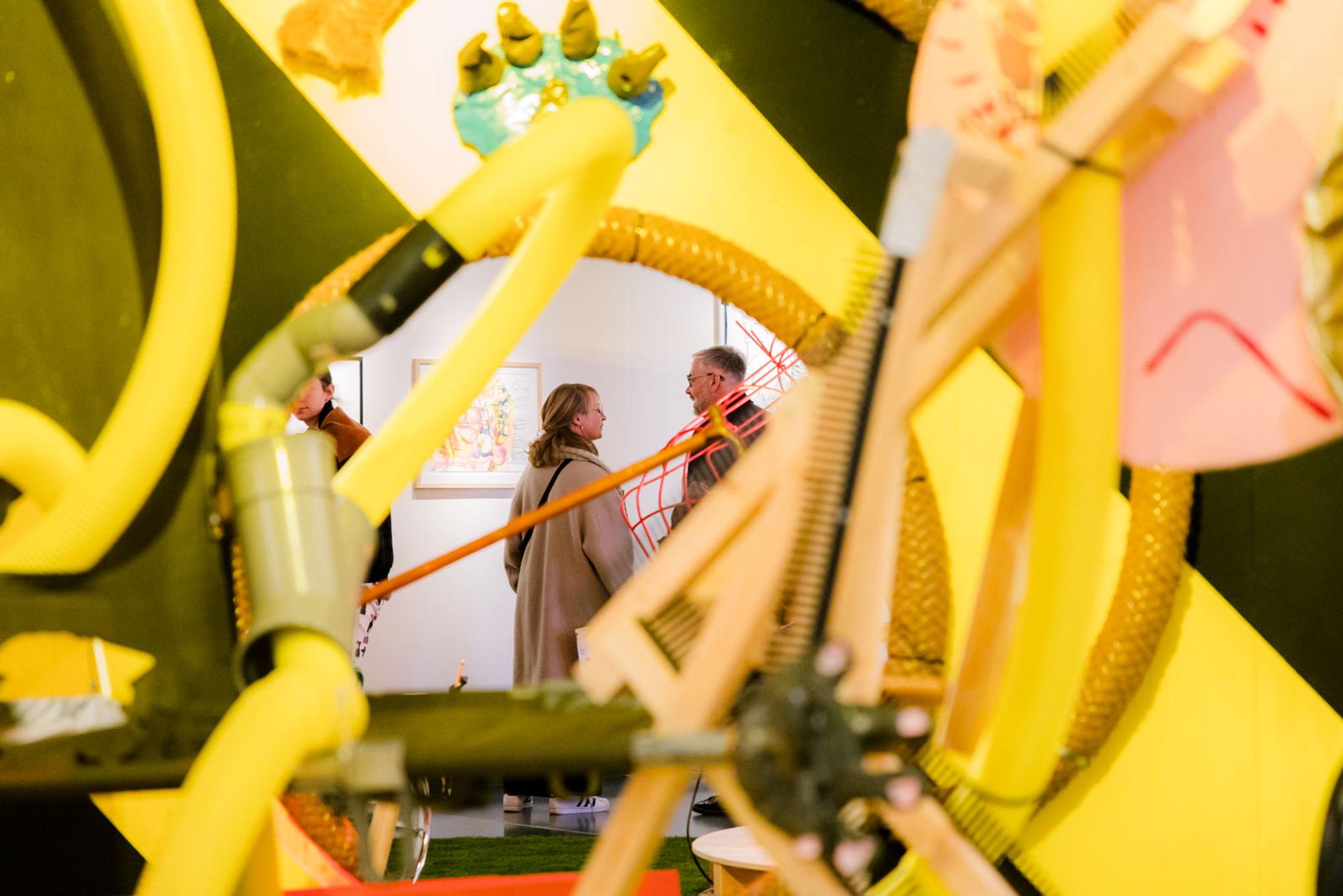 Kunstwerk met gele buizen met man en vrouw op de achtergrond die met elkaar praten. 