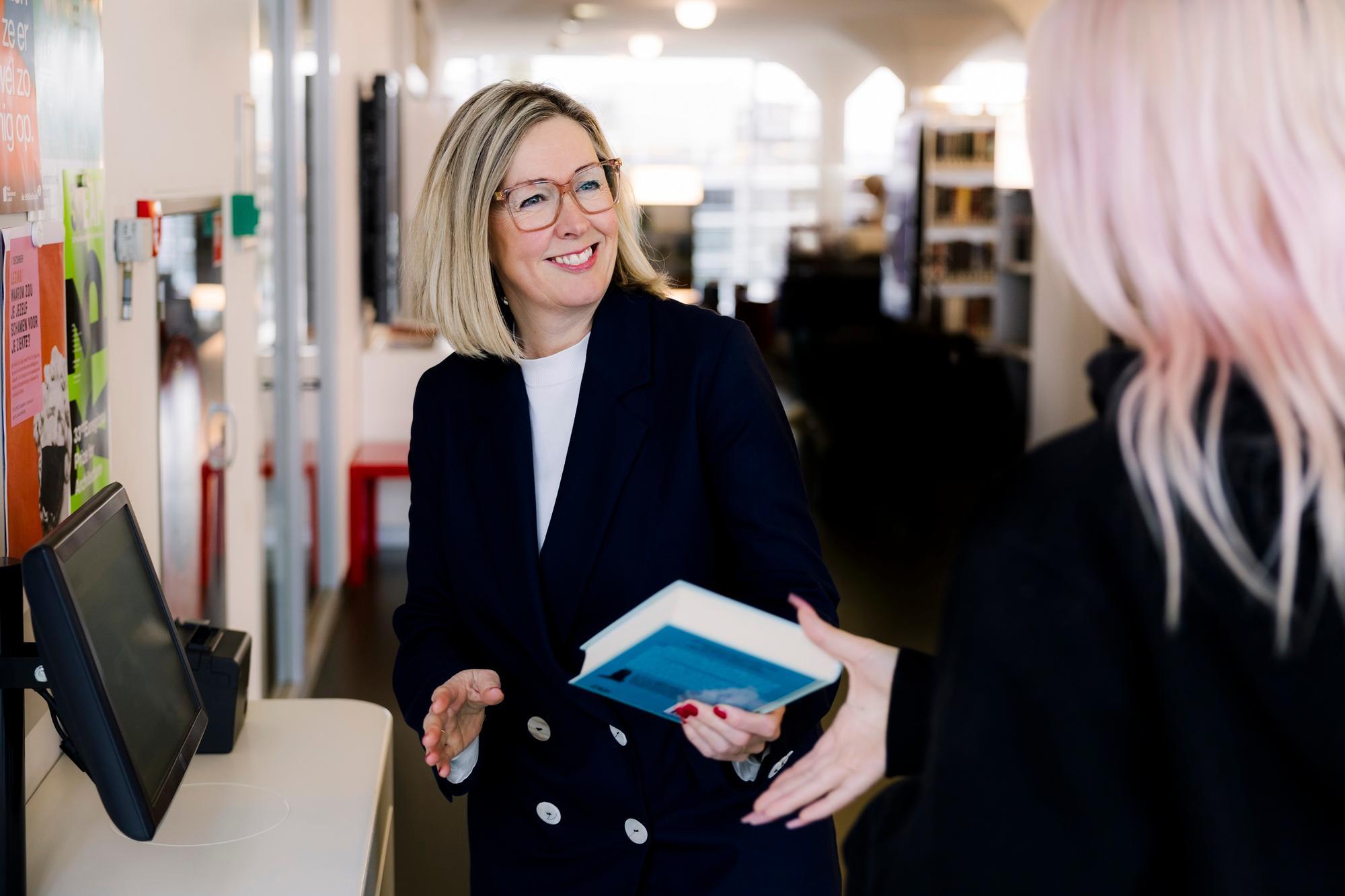 Vrouw met bril overhandigt een boek aan een bezoeker in de bibliotheek.