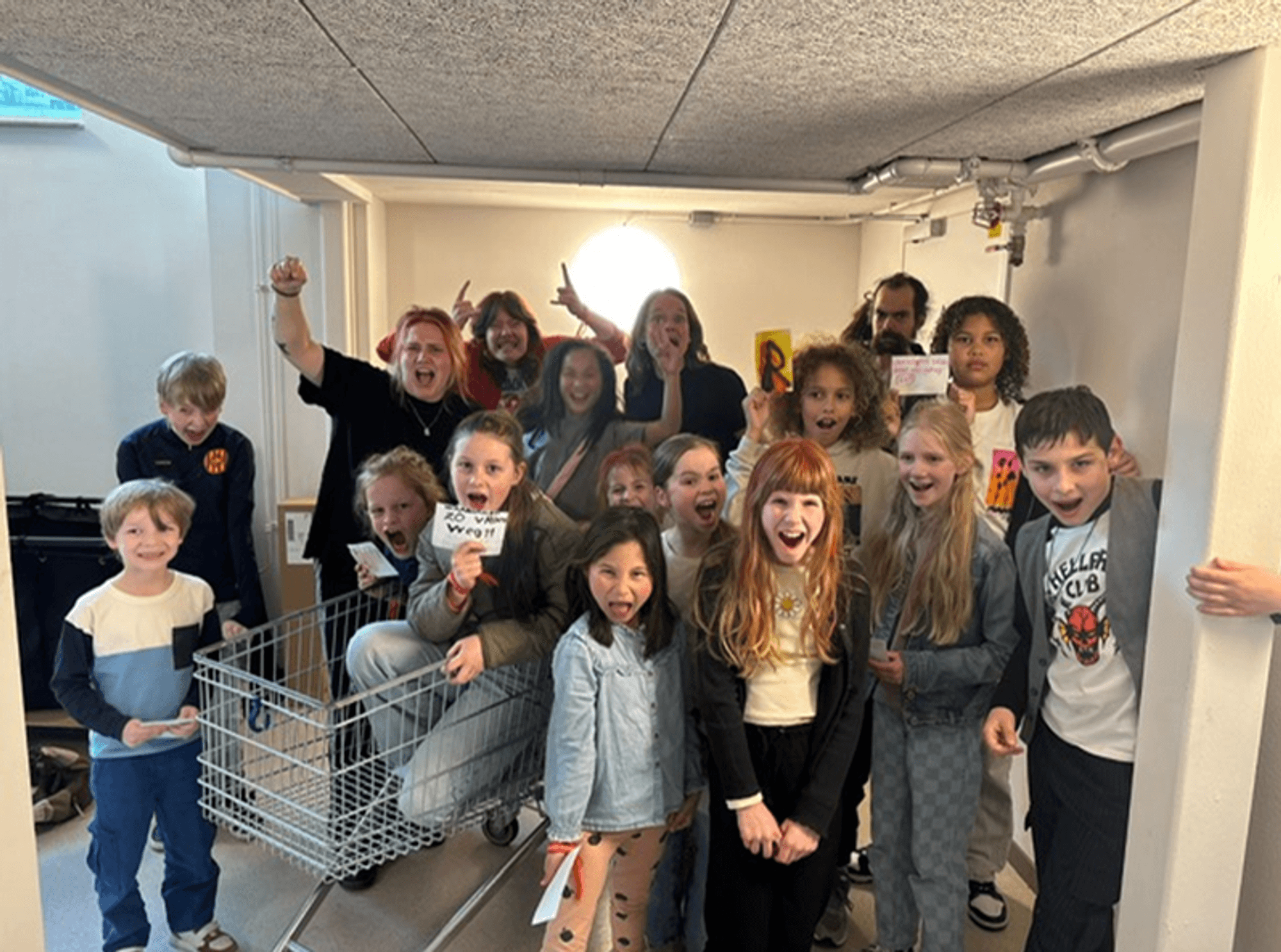 Groep kinderen en volwassenen poseert in een ruimte rond een winkelwagentje en houdt kaartjes vast tijdens een activiteit binnenshuis.
