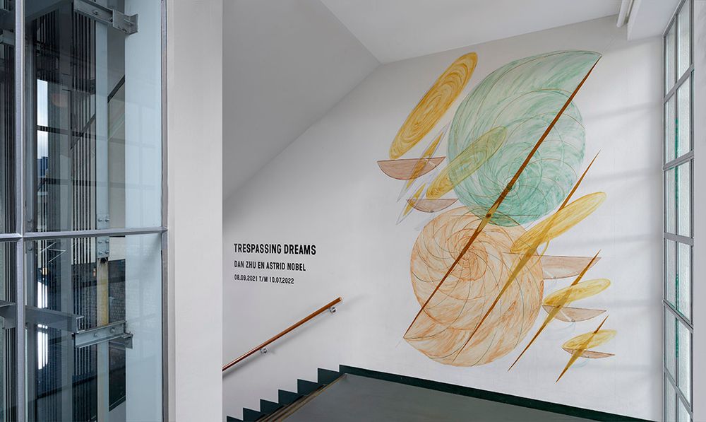 Muurschildering in het trappenhuis van SCHUNCK met abstracte, cirkelvormige figuren in pastelkleuren en schuine lijnen, naast de titel Trespassing Dreams van Dan Zhu en Astrid Nobel.