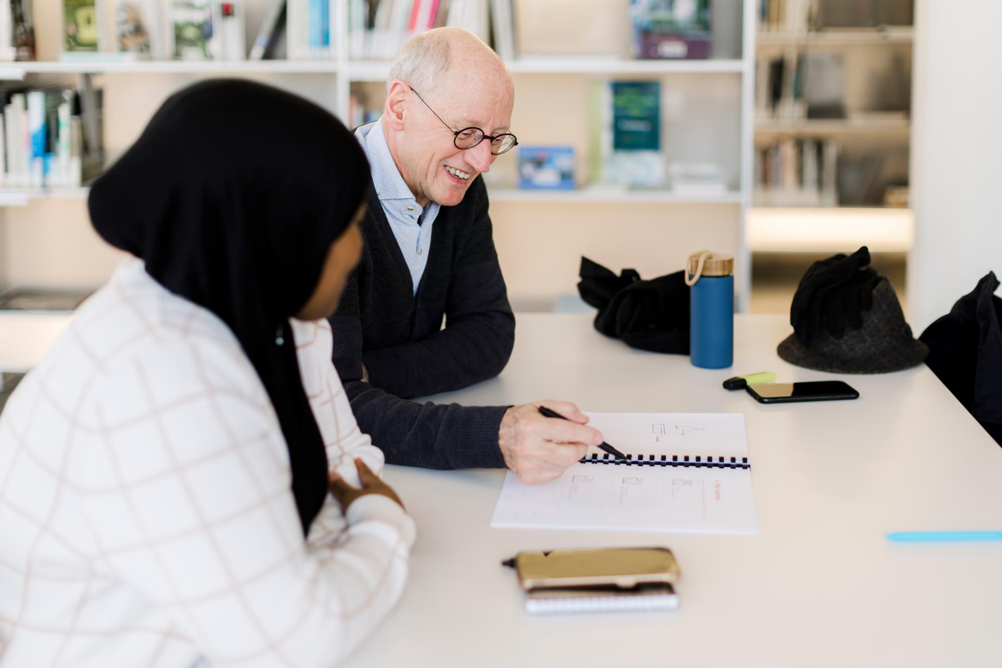 Man en vrijwilliger zitten aan een tafel in de bibliotheek. De man wijst met een pen naar een papier voor hem op tafel.