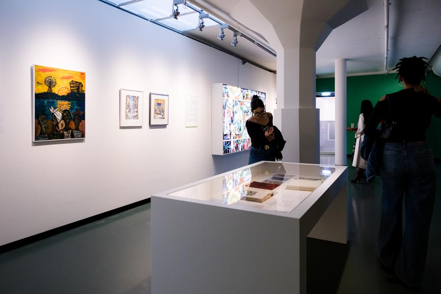 Bezoekers bekijken kunstwerken in een tentoonstellingsruimte van SCHUNCK Museum. Aan de muur hangen kleurrijke schilderijen en tekeningen; in het midden staat een vitrinetafel met archiefmateriaal. Op de achtergrond is een groen geschilderde muur zichtbaar.