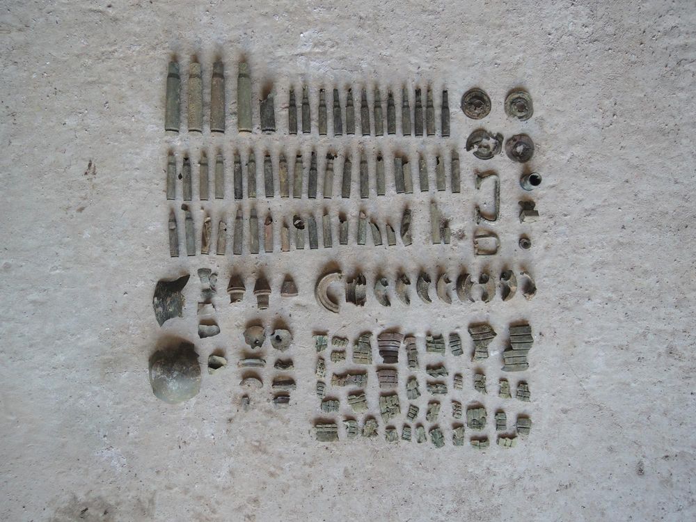 Een verzameling oude metalen objecten, waaronder hulzen, ringen en fragmenten, ligt geordend in strakke rijen op een lichte ondergrond. De stukken zijn verweerd en doen denken aan gevonden resten van munitie of metaal.