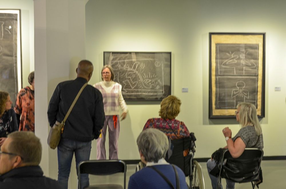 Een groep mensen in een kunstgalerij. Verscheidene individuen zitten, terwijl anderen staan en de tentoongestelde kunstwerken bekijken. De kunstwerken lijken ingelijste stukken met abstracte ontwerpen of tekeningen te zijn.