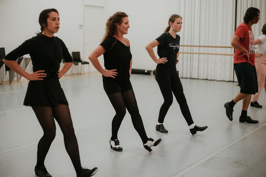 Drie vrouwen en een man dansen met hun handen in de zij en een been naar voren, waarbij hun hak op de grond staat.