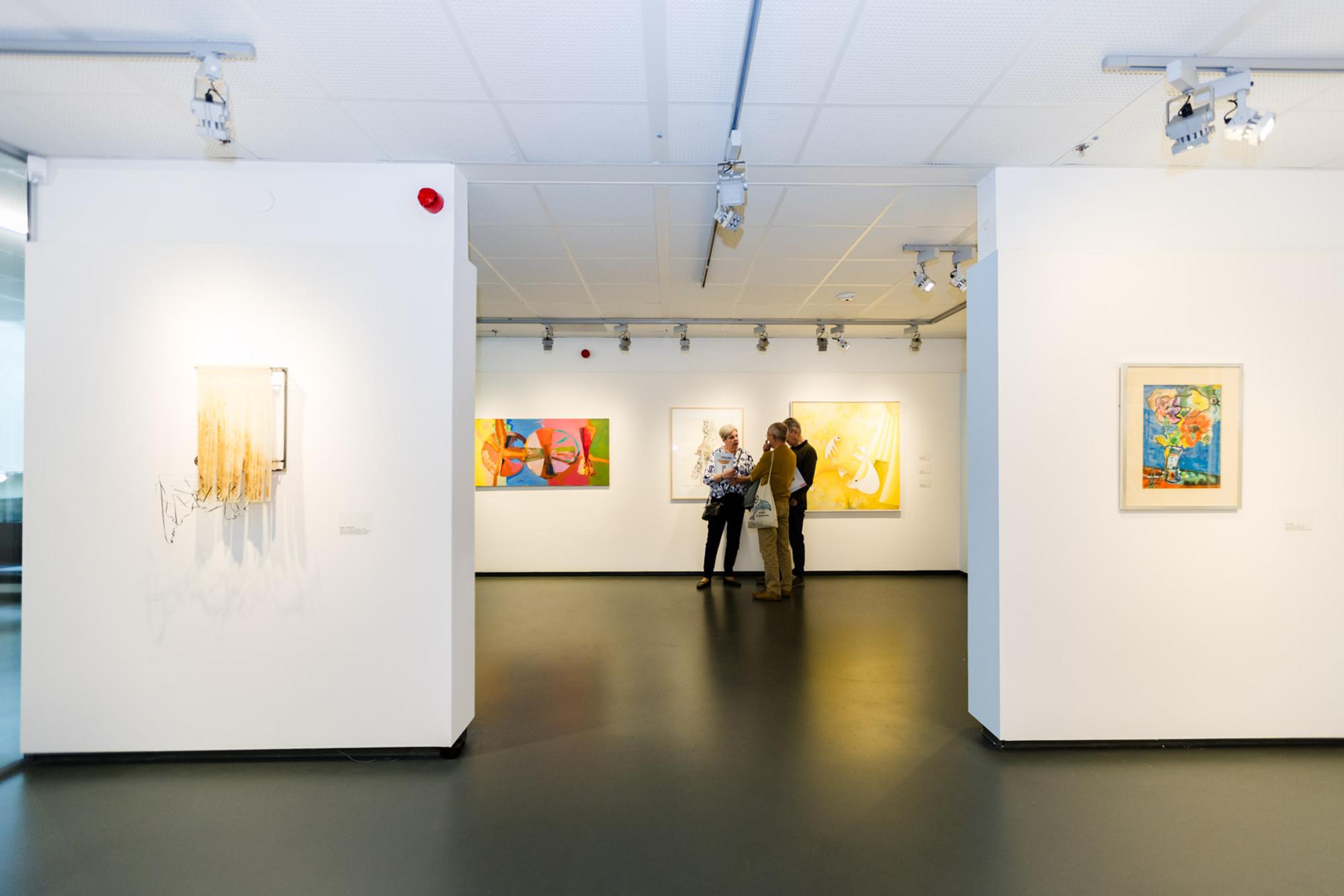 Drie bezoekers kijken naar kunst in Aad de Haas zaal. Kunstwerken hangen aan de muur. 