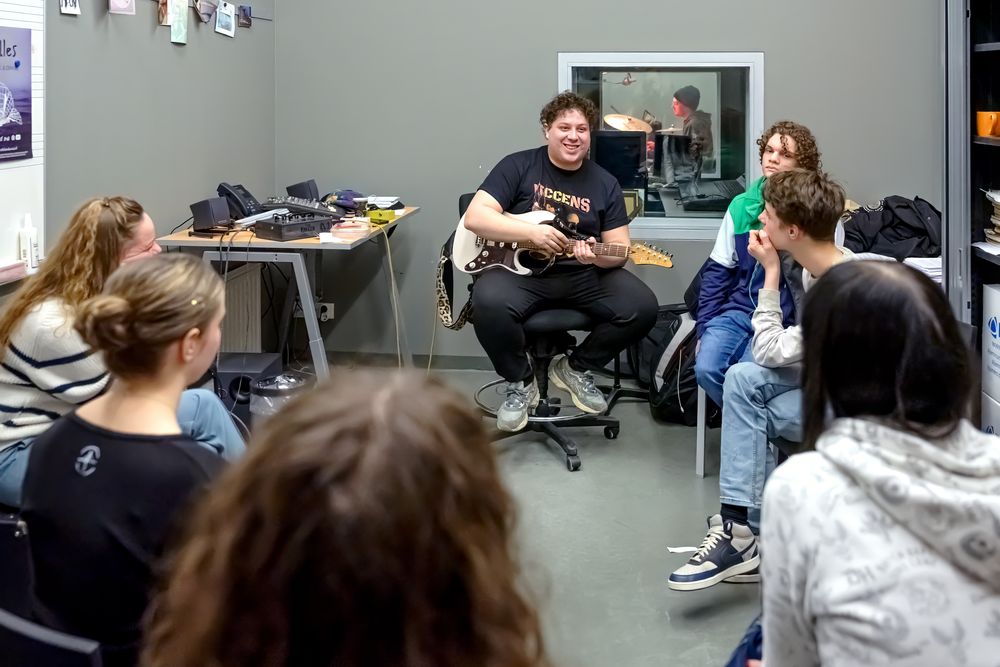Zeven jongeren zitten in een cirkel in een muzieklokaal. Een jongen met een gitaar kijkt vrolijk terwijl de anderen naar hem kijken.