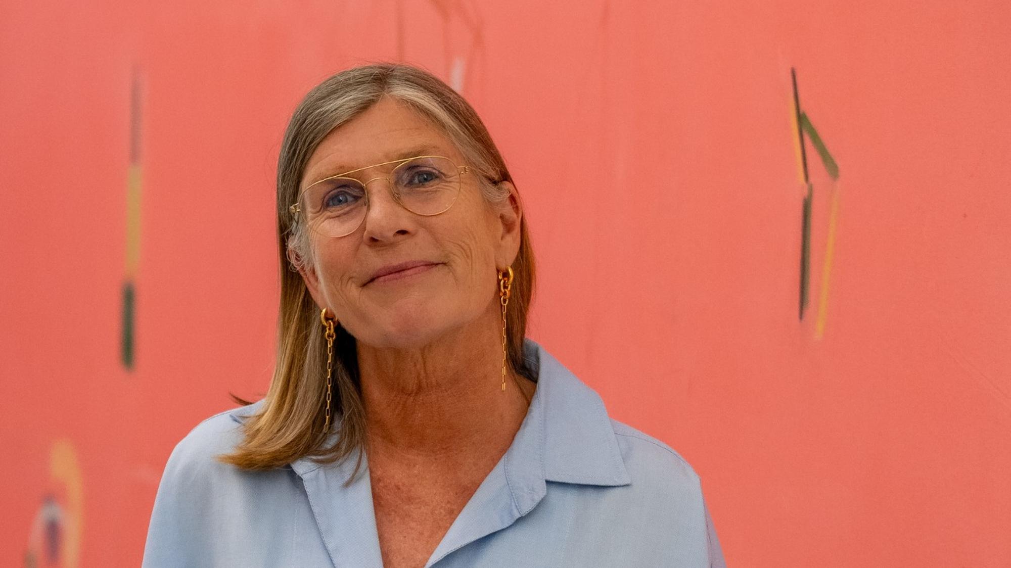 Portret van Catharien Romijn in een lichtblauwe blouse, volledig in beeld, staand voor een roze kunstwerk met subtiele abstracte vormen.