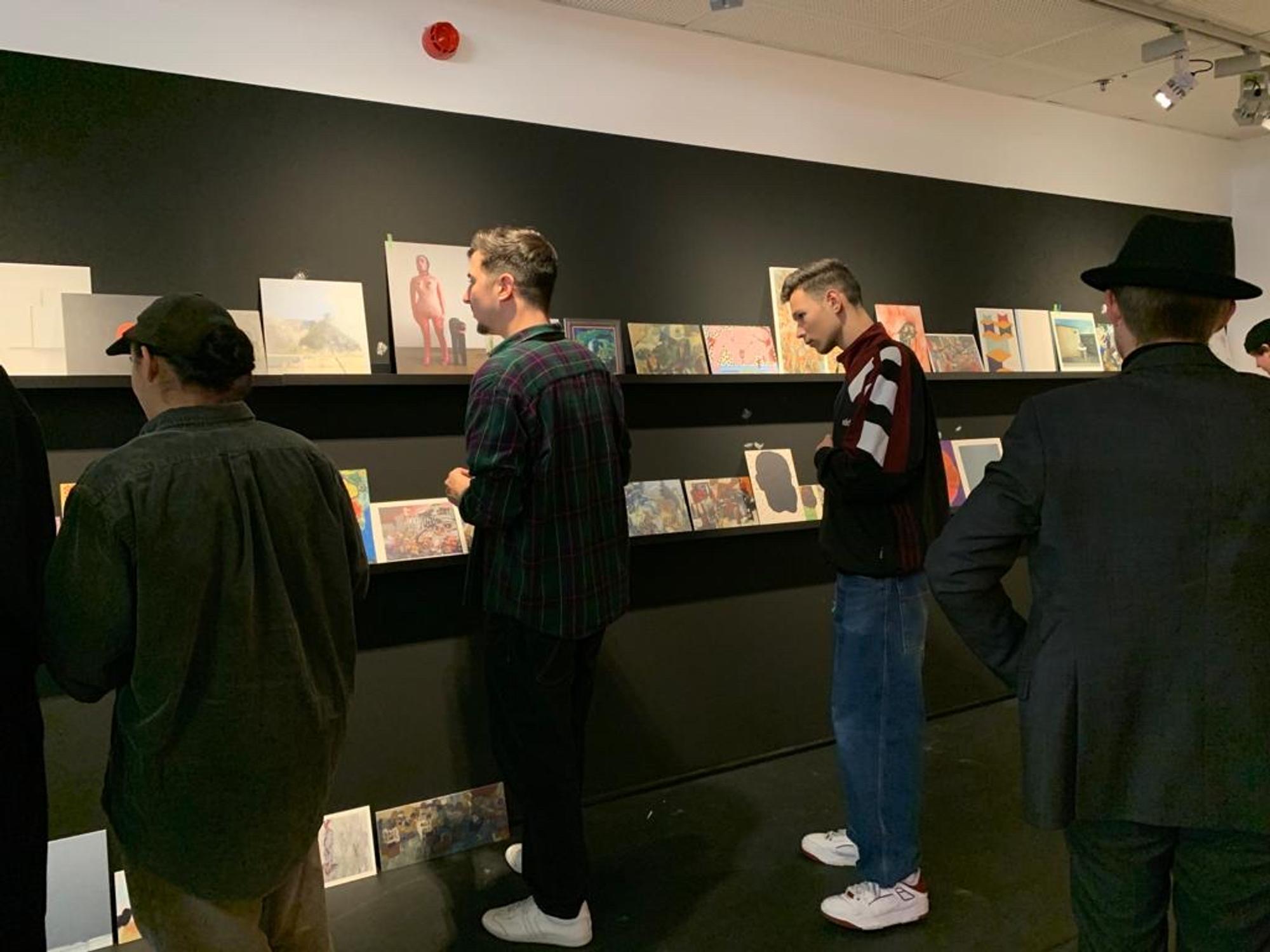drie mannen kijken naar verschillende kunstwerken tegen een zwarte muur.