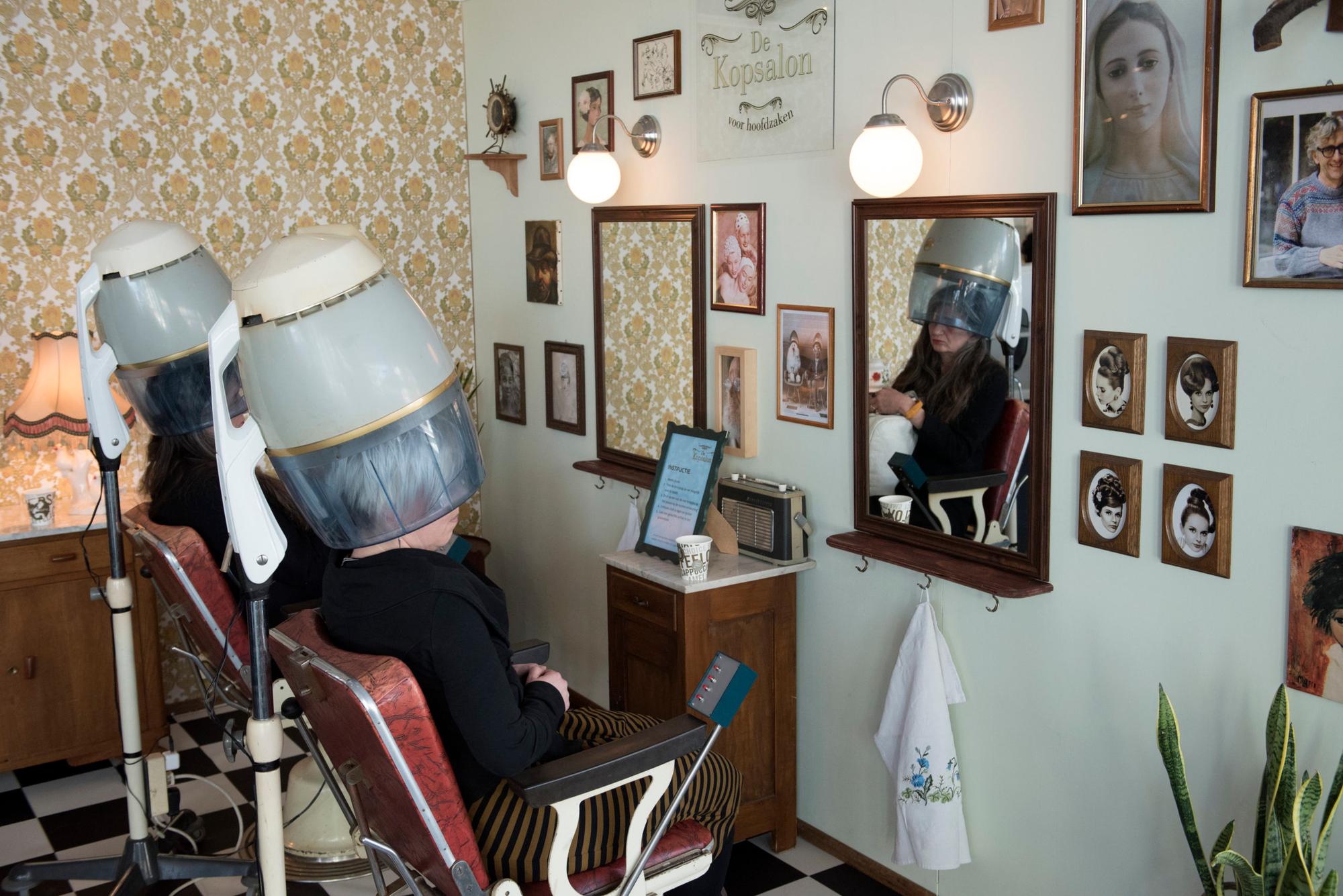 Een wat oudere foto van een kapsalon waarbij twee vrouwen met kappen op hun hoofd zitten.