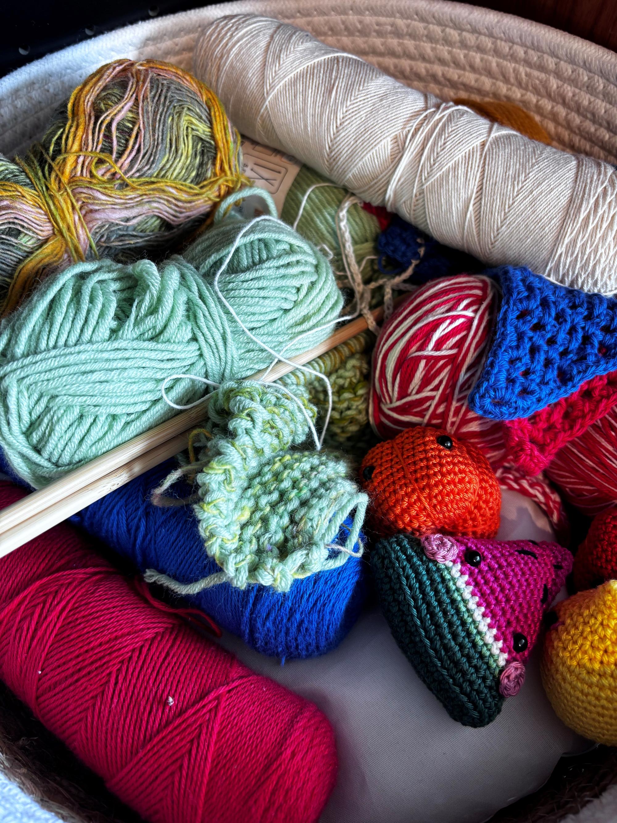 Close-up van een mand met gekleurde bollen wol, breinaalden en gehaakte figuurtjes in de vorm van fruit. De materialen liggen door elkaar, wat een creatief en ambachtelijk geheel vormt.