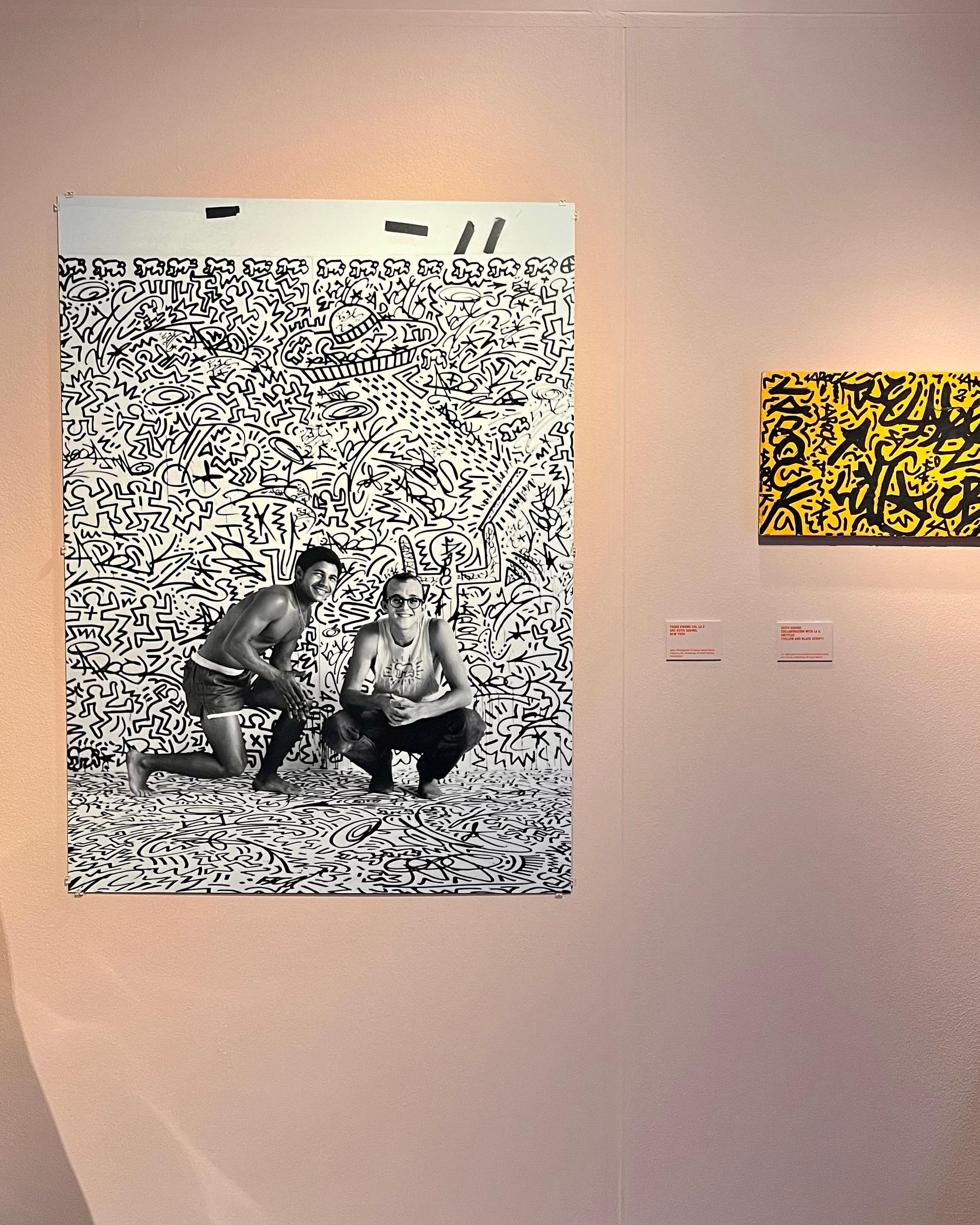 Zwart-witfoto van twee lachende personen zittend en hurkend voor een met Keith Haring-illustraties bedekte muur en vloer. Rechts hangt een felgeel schilderij met zwarte graffiti-achtige figuren in dezelfde expressieve stijl.