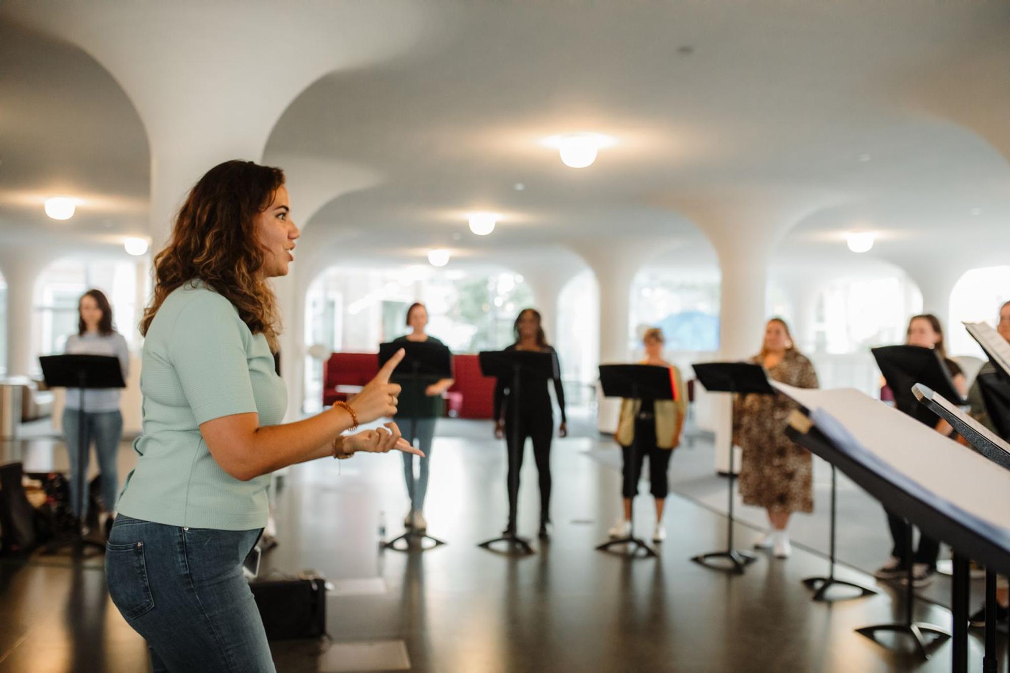 Een groep mensen staat in een cirkel met elk een muziekstandaard en zingen. In het midden staat de docent die met haar handen een gebaar maakt.
