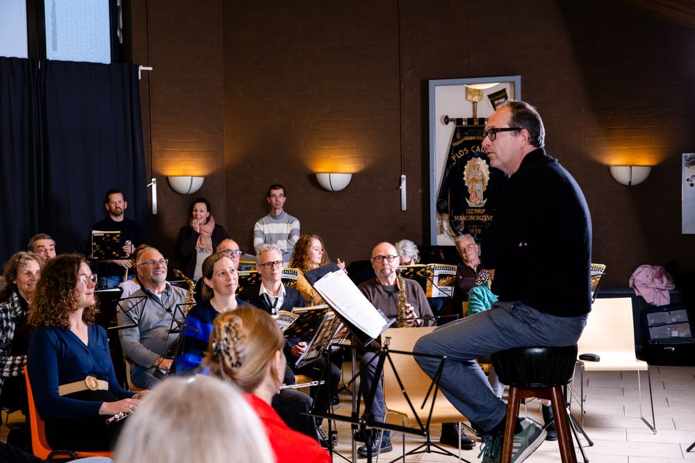 Een dirigent zit voor een groep volwassen muzikanten tijdens een repetitie en geeft uitleg of instructies. De deelnemers luisteren aandachtig met hun instrumenten en bladmuziek voor zich, in een ontspannen en betrokken sfeer.