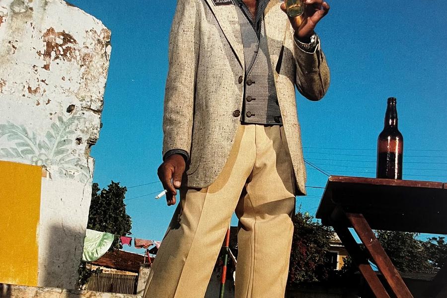 Fotografisch portret van een man die buiten staat op straatstenen, gekleed in een colbert en lichte broek, met een glas in zijn hand tegen een blauwe lucht.