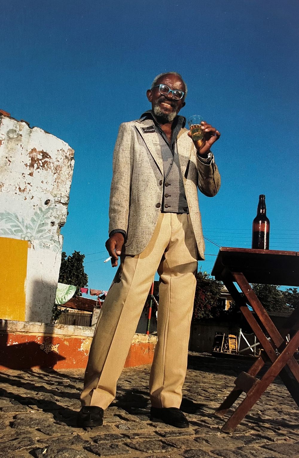 Fotografisch portret van een man die buiten staat op straatstenen, gekleed in een colbert en lichte broek, met een glas in zijn hand tegen een blauwe lucht.