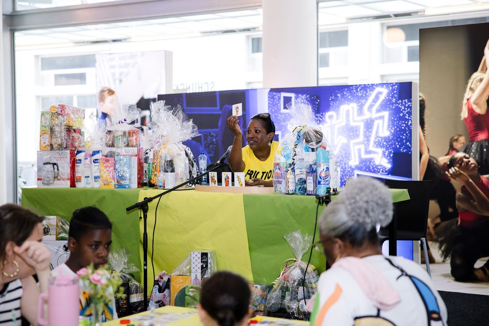 Een vrouw in een geel T‑shirt zit achter een tafel met groene en gele doeken, houdt een kaart omhoog en presenteert prijzen tijdens het spel Bon Ku Ne op Frictions Festival #9 bij SCHUNCK; op de tafel staan ingepakte cadeaus en verzorgingsproducten, terwijl bezoekers aan tafels met bloemen en drankjes aandachtig meespelen.