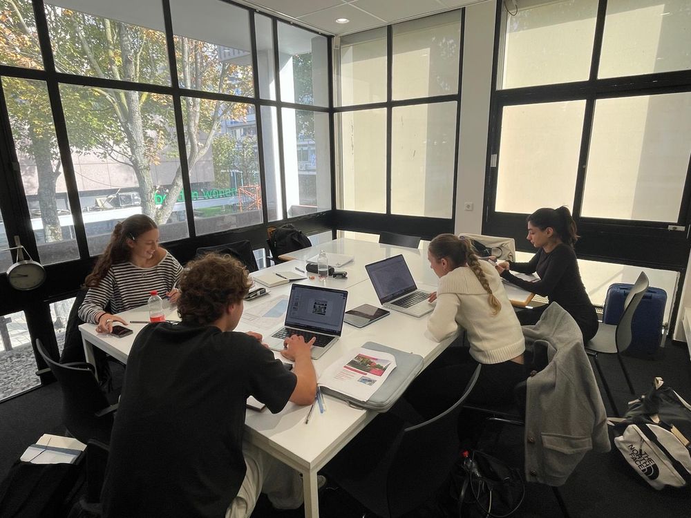 Vier studenten werken aan een grote witte tafel met laptops, tekeningen en documenten tijdens een groepssessie voor de Euregionale Architectuurprijs.