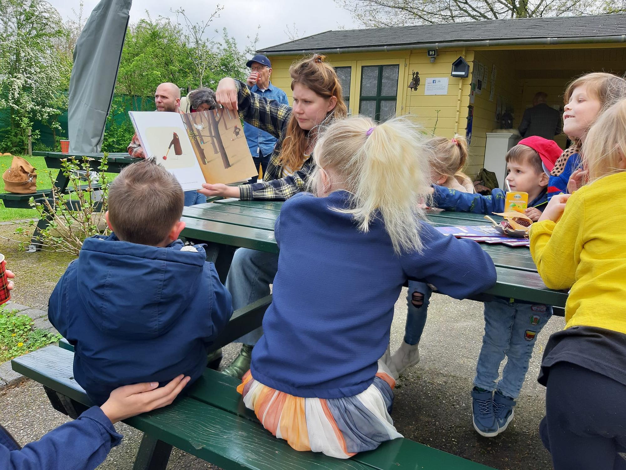 Kinderen luisteren buiten naar een voorlezer die een boek toont bij een groene picknicktafel, met een geel gebouw en bomen op de achtergrond.