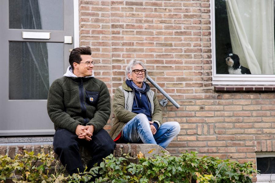 Jop Vermeesch en zijn vader zitten samen op de stoep voor hun woning, met een hond die door het raam meekijkt.