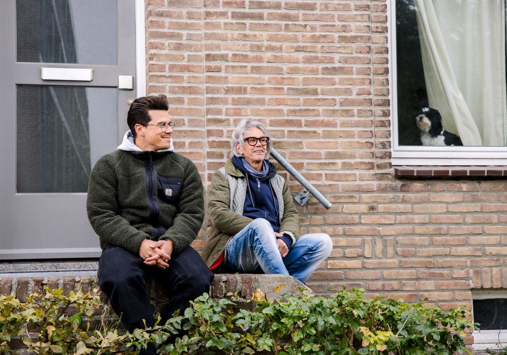 Jop Vermeesch en zijn vader zitten samen op de stoep voor hun woning, met een hond die door het raam meekijkt.