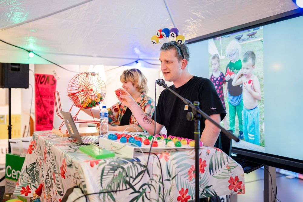 Kunstenaar Maarten Bel presenteert een vrolijke bingo‑sessie onder een tent tijdens Frictions Festival #12; hij draagt een hoofdband met biljartballen, spreekt in een microfoon en houdt een gekleurde bingobal omhoog, terwijl achter hem een groot scherm een foto van kinderen toont en een assistent naast een rode bingomolen zit aan een tafel met bloemenpatroon.