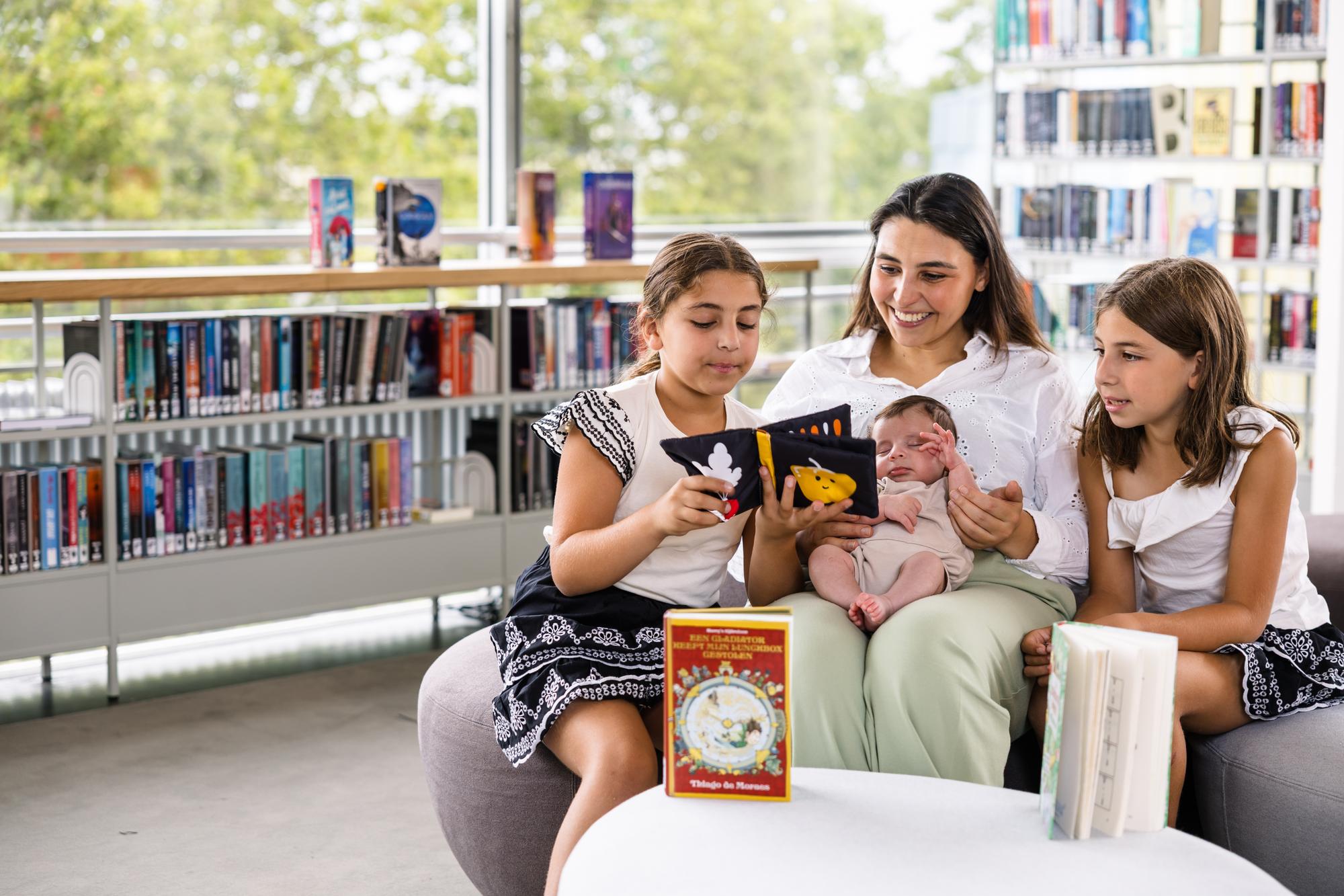 Een groep kinderen en een volwassene zitten samen in een bibliotheek en bekijken een boek, terwijl een baby op schoot wordt vastgehouden. Op de achtergrond zijn boekenkasten en grote ramen met uitzicht op groen te zien.