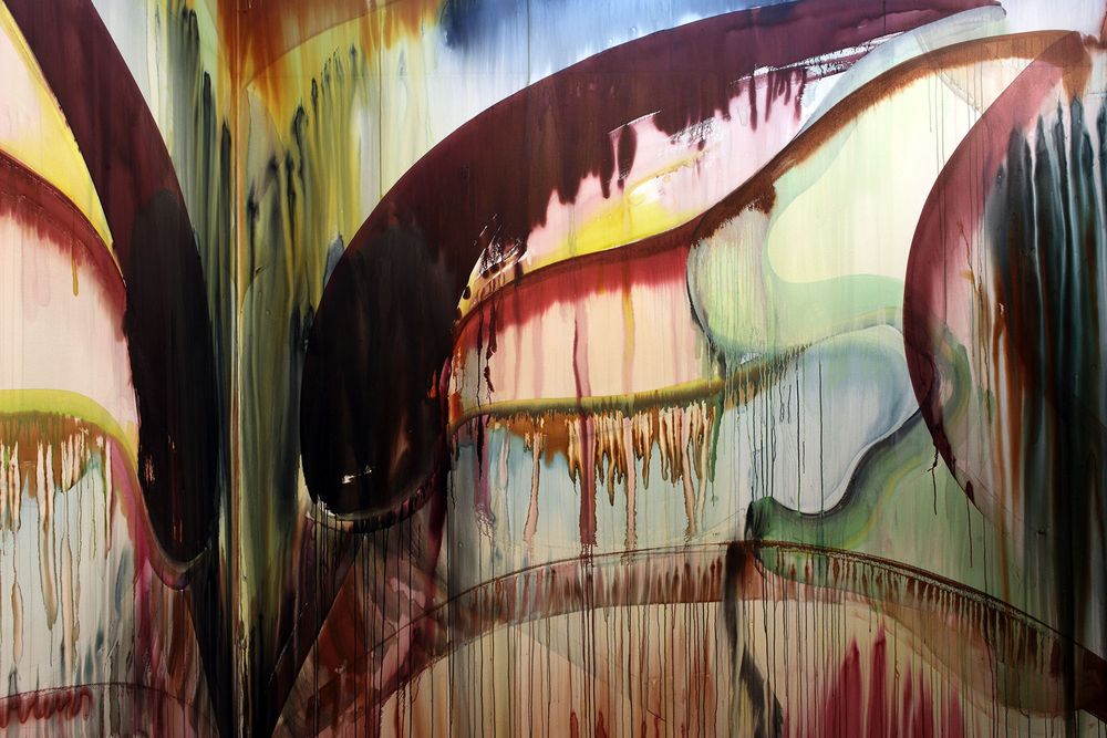 Groot abstract schilderij met druipende verfbanen in rood, geel, zwart, groen en blauw, die over elkaar heen vloeien op een wand in een tentoonstellingsruimte.