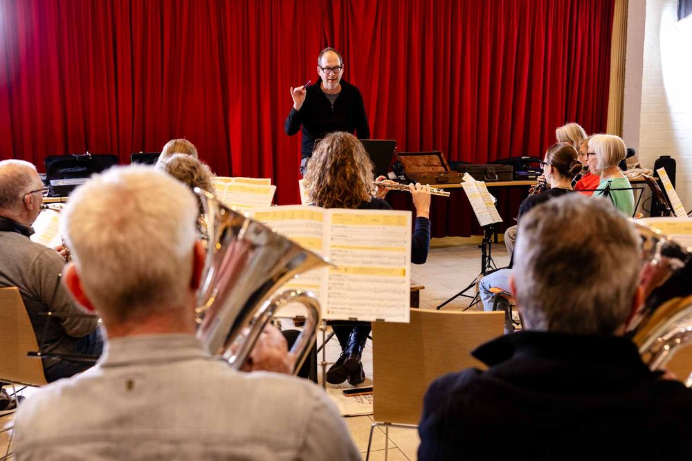 Dirigent leidt een harmonieorkest tijdens een repetitie, terwijl de muzikanten met bladmuziek voor zich spelen. Op de achtergrond hangt een rood gordijn dat het podium sfeervol omlijst.