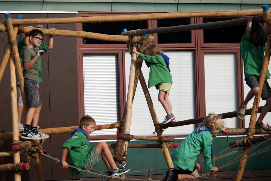 Kinderen klimmen en balanceren op een speelconstructie van houten palen en touwen buiten bij een gebouw.
