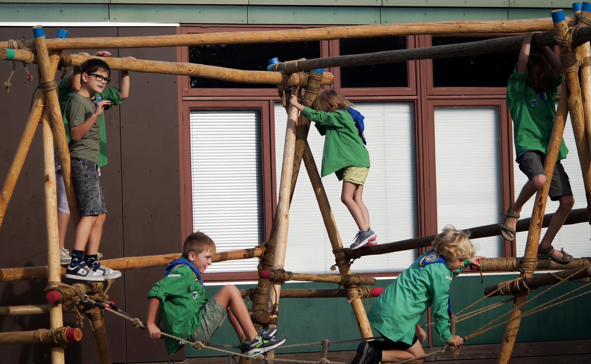 Kinderen klimmen en balanceren op een speelconstructie van houten palen en touwen buiten bij een gebouw.