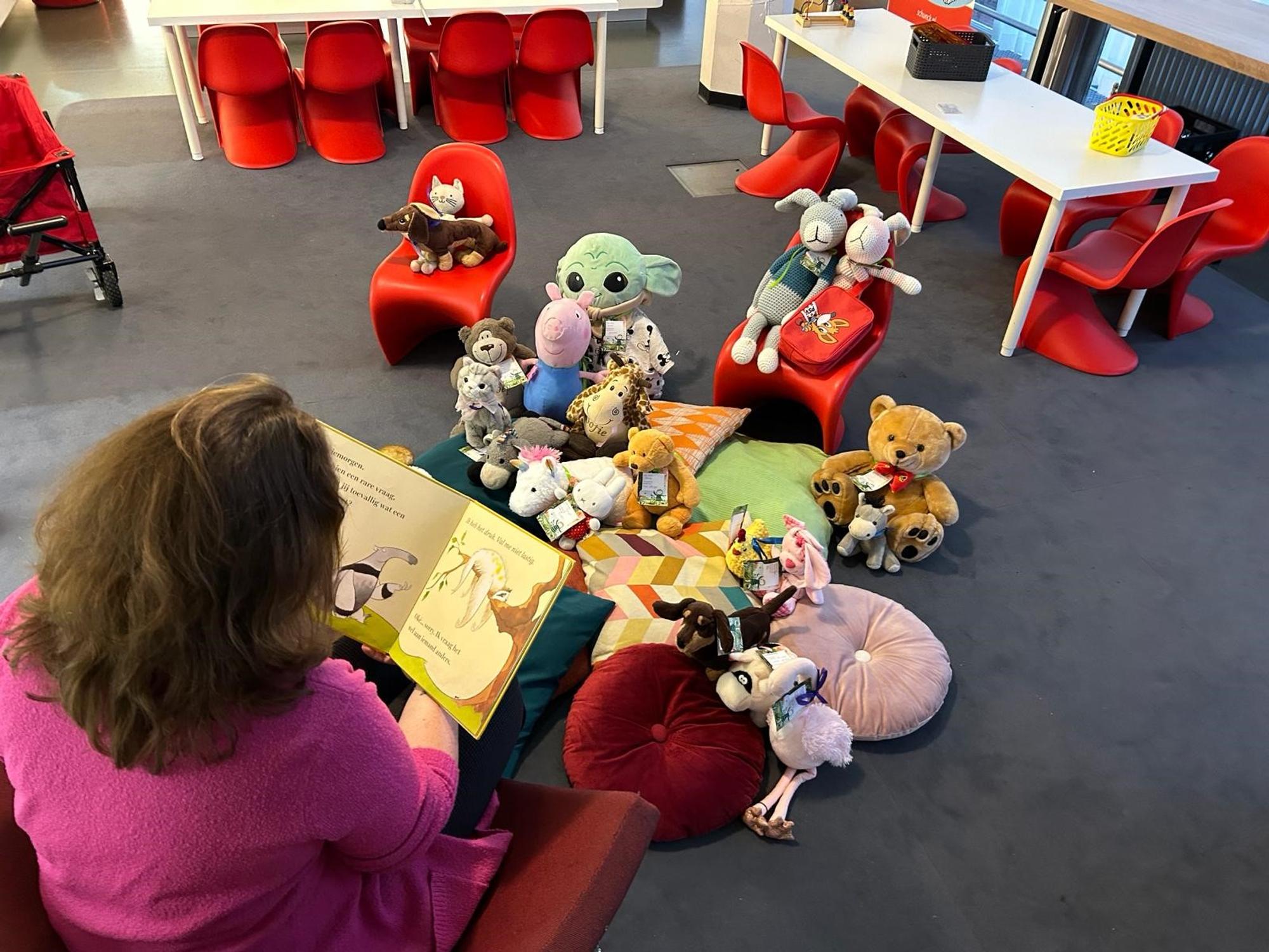 Vrouw in een roze trui leest voor uit een prentenboek aan een kring van knuffeldieren die op kussens en stoeltjes in een kinderruimte zitten, met rode stoelen en tafels op de achtergrond.