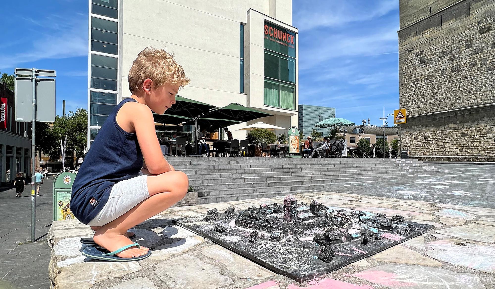 Kind zit buiten in de zon naast een maquette van Heerlen, voor het SCHUNCK Glaspaleis.