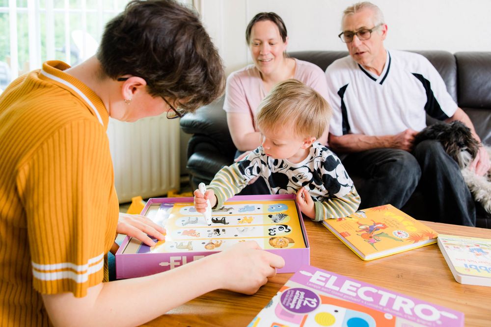 Jong kind. tekent met een dikke pen terwijl naast hem twee volwassenen vrolijk toekijken. 