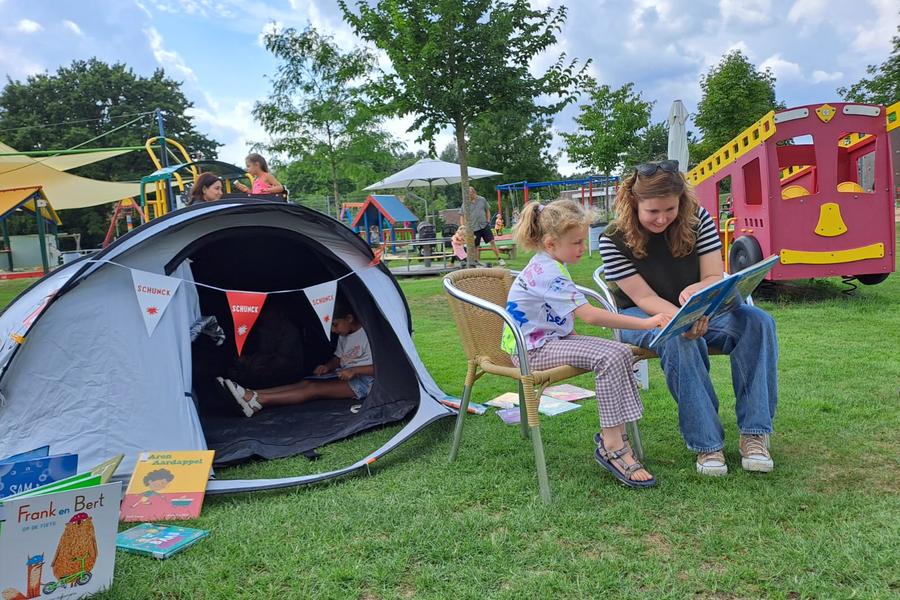 Tijdens een voorleescamping in een speeltuin leest een vrouw samen met een jong meisje een prentenboek op het gras. Naast hen staat een tent waarin kinderen lezen, terwijl speeltoestellen en andere bezoekers op de achtergrond zorgen voor een gezellige buitensfeer.
