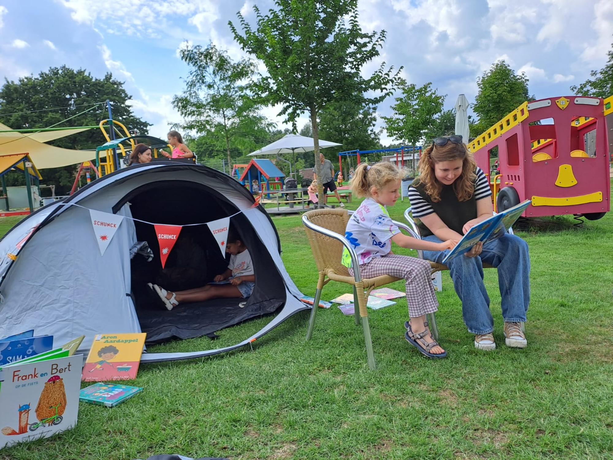 Tijdens een voorleescamping in een speeltuin leest een vrouw samen met een jong meisje een prentenboek op het gras. Naast hen staat een tent waarin kinderen lezen, terwijl speeltoestellen en andere bezoekers op de achtergrond zorgen voor een gezellige buitensfeer.
