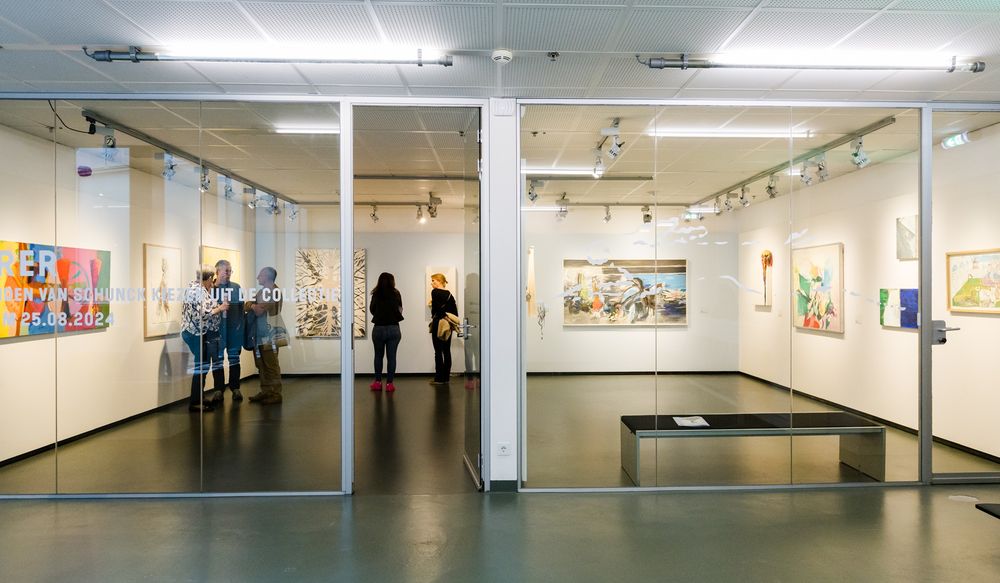 Expositie met schilderijen uit de collectie van SCHUNCK in de Aad de Haas zaal: een ruimte met grote  ramen. Bezoekers staan in de ruimte en kijken naar de kunstwerken.