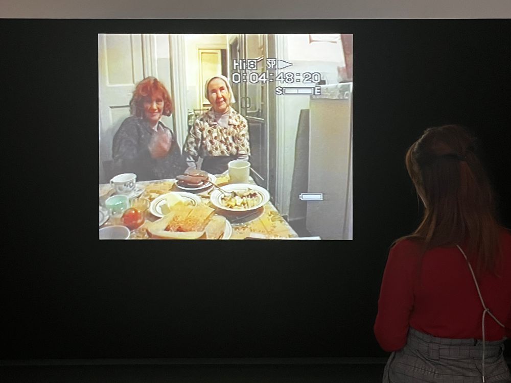 Een vrouw bekijkt een videoprojectie op een donkere muur. Op het scherm zitten twee vrouwen aan een gedekte keukentafel.