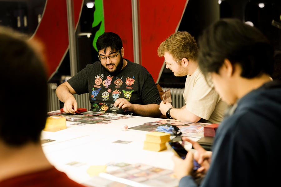 Een groep mensen zit samen aan een tafel in een fel verlichte ruimte terwijl ze een kaartspel spelen. Op de tafel liggen speelmatten, stapels kaarten en dobbelstenen. De sfeer oogt ontspannen en geconcentreerd.