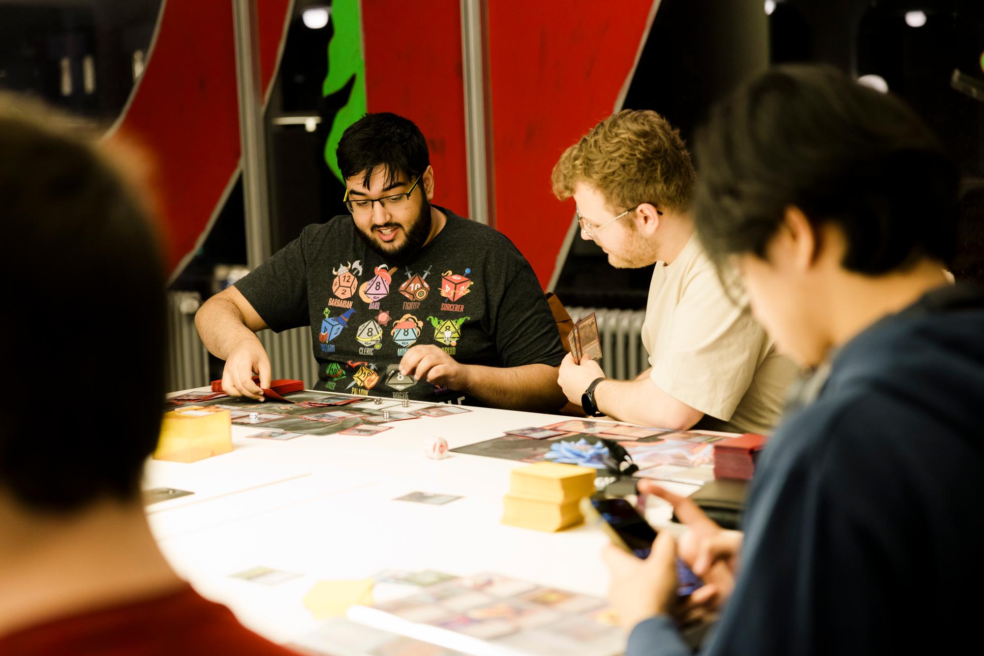 Een groep mensen zit samen aan een tafel in een fel verlichte ruimte terwijl ze een kaartspel spelen. Op de tafel liggen speelmatten, stapels kaarten en dobbelstenen. De sfeer oogt ontspannen en geconcentreerd.