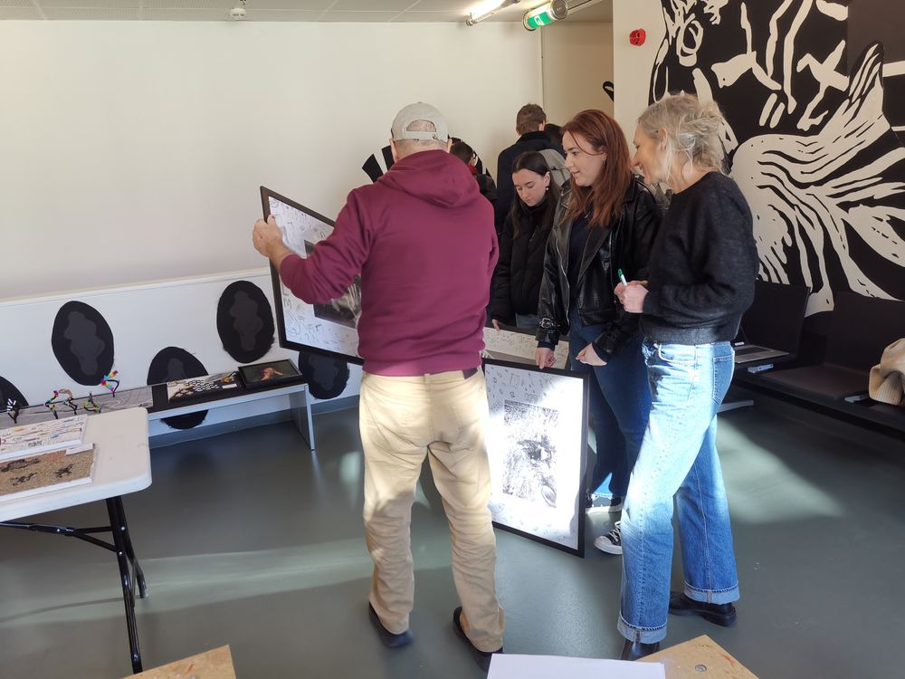 Groep mensen bespreekt ingelijste kunstwerken in een expositieruimte met zwart-witte muurschilderingen; een man in hoodie en pet houdt een lijst vast terwijl anderen aandachtig kijken.