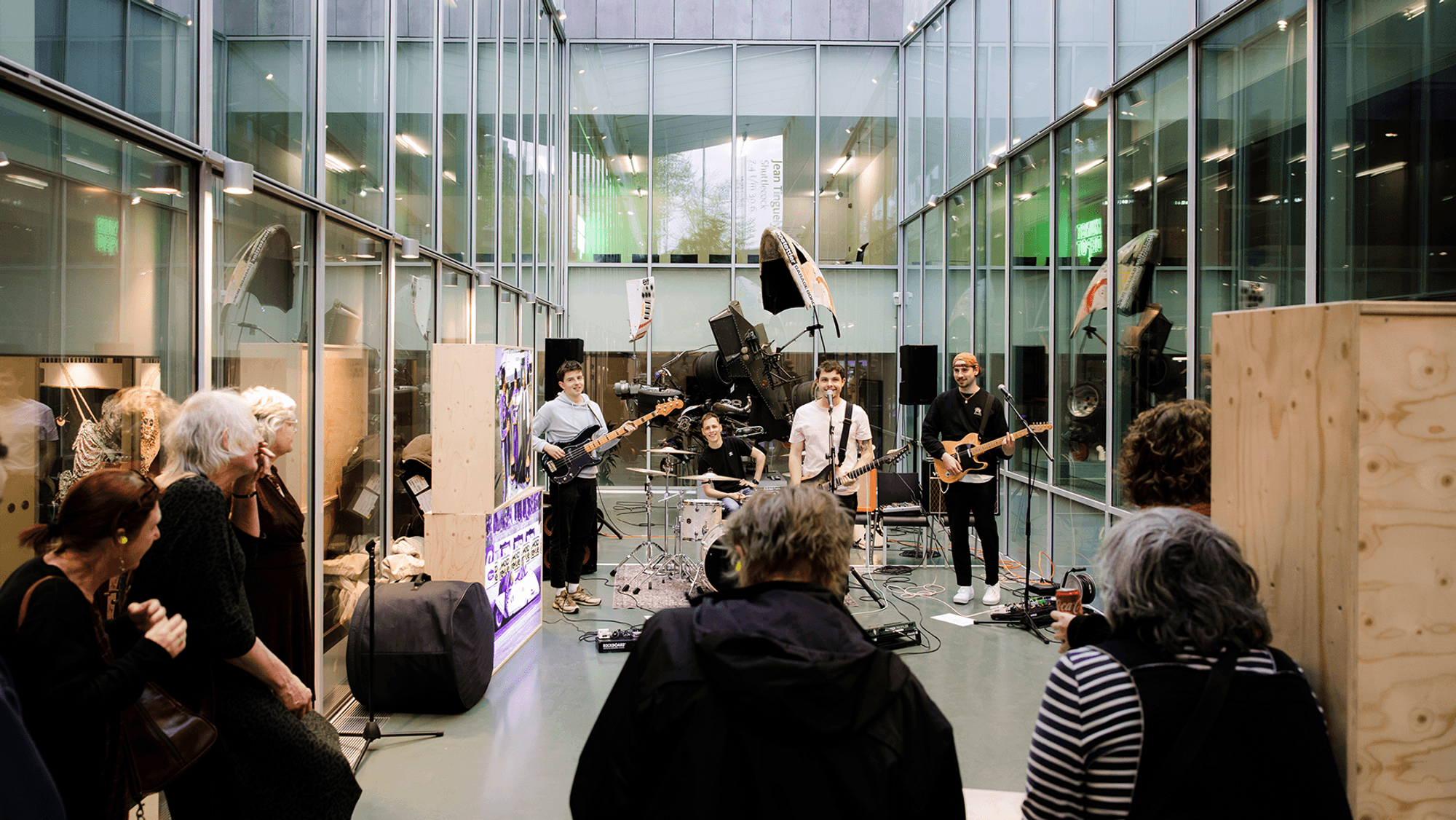 Bandje met vier jonge mannen treden op in het Atrium. Drie jongens spelen gitaar en een drumt. Op de achtergrond is een installatie van Jean Tinguely te zien. Op de voorgrond zijn bezoekers te zien.