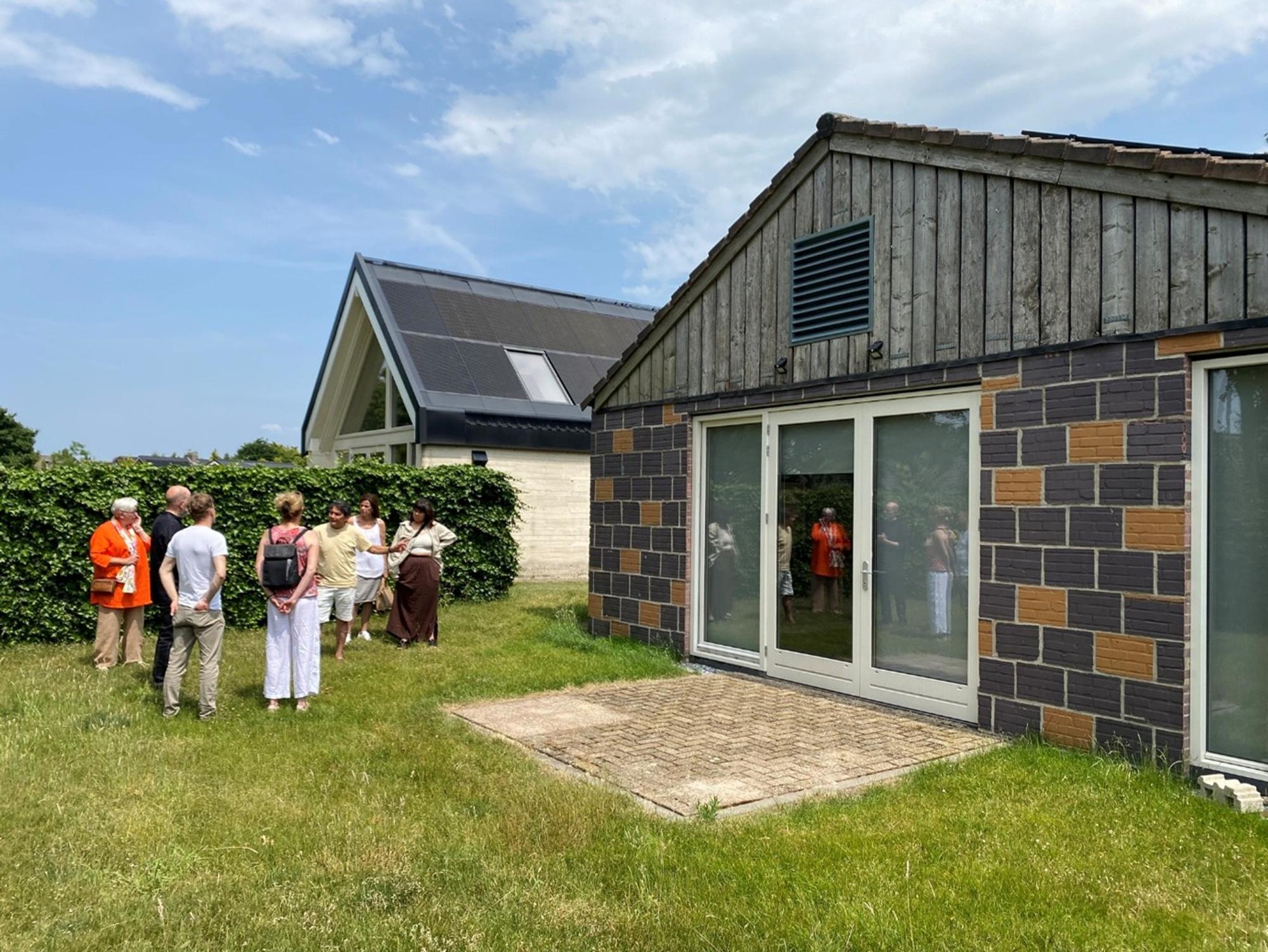 Groep mensen staat in een tuin naast een modern gebouw tijdens een rondleiding langs woningen met verschillende architectuurstijlen.